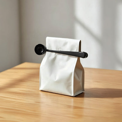 Black EspressoWorks coffee scoop clipped onto a white bag of coffee on a wooden table