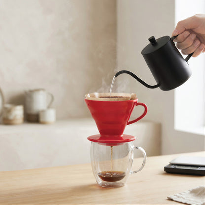 Pouring hot water from a gooseneck kettle into the red pour over dripper on a kitchen counter
